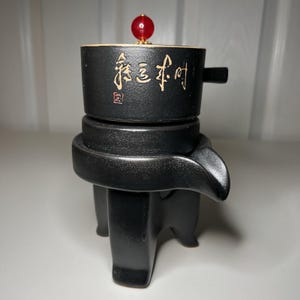 May include: Black ceramic tea infuser with a three-legged base and a spout. The infuser has a cylindrical body with gold Chinese characters and a red bead on top.