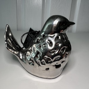 May include: A silver ceramic bird figurine with a glossy finish. The bird has a detailed design with cut-out patterns on its body. It has a small beak and a tag attached to it. The figurine is sitting on a white surface.