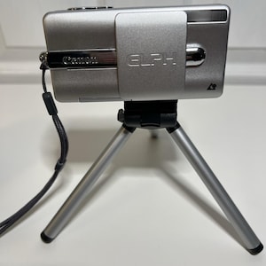 May include: A silver Canon ELPH digital camera mounted on a small, silver tripod. The camera has a black wrist strap attached. The tripod has three legs and is designed for stability. The camera is in a horizontal position.