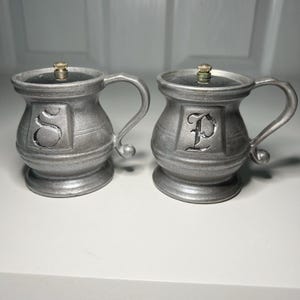 Könnte beinhalten: Zwei salz- und pfefferstreuer in Zinnfarbe. Jeder Shaker hat die Form eines kleinen Bechers mit Henkel und Deckel. Ein Shaker hat ein "S" und der andere ein "P" auf der Vorderseite. Die Deckel haben einen kleinen goldfarbenen Knopf.