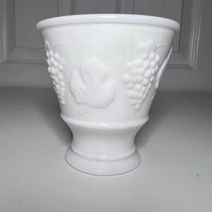 May include: A white, vintage-style vase with a flared top and a pedestal base. The vase features embossed grape and leaf designs around the exterior. The vase is made of opaque white glass and is suitable for floral arrangements or decorative use.