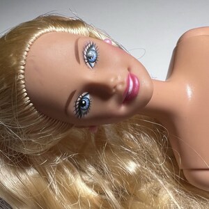 May include: A doll's head with blonde hair and blue eyes. The doll has pink lips and a fair complexion. The head is tilted, and the hair is flowing. The doll's eyes are detailed with black eyeliner and eyelashes.