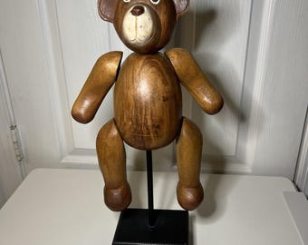 Puede incluir: Figura de oso de peluche de madera con extremidades articuladas, montada sobre un soporte negro. El oso tiene la cara y las orejas de color marrón claro, con vetas de madera marrón oscuro en el cuerpo y las extremidades. Los ojos son blancos con pupilas negras.