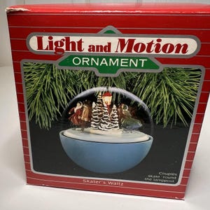 Peut inclure: Boîte rouge avec l'inscription « Light and Motion Ornament » et le texte « Skater's Waltz ». À l'intérieur, une base bleue en forme de bol avec un dôme transparent représentant une scène hivernale.