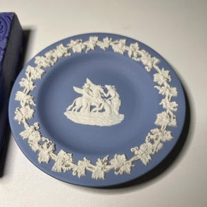 May include: A blue decorative plate with a white relief design. The plate features a central scene of figures and a border of grapevines. The plate is round and appears to be made of ceramic.