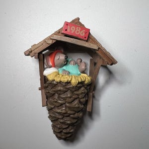 以下が含まれることがあります： 松ぼっくりの家で眠っているネズミを描いた、ヴィンテージのクリスマスオーナメント。 ネズミは赤い帽子と青いパジャマを着て、黄色のわらの上に休んでいます。 オーナメントは茶色で、「1986」と書かれた赤いサインが付いています。