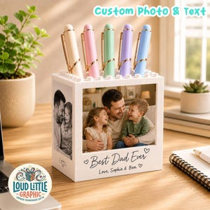 May include: A white block-style desk organizer featuring a custom photo of a father and two children. The front displays the text "Best Dad Ever" and "Love, Sophie & Ben" with heart icons. Five pastel-colored pens are stored in the top slots.