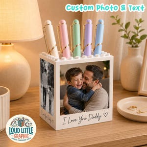 May include: A white square desk organizer featuring a custom photo of a child and adult on the front, with the text I Love You Daddy and a heart. The top holds five pens in cream, pink, light green, lavender, and light blue colors.