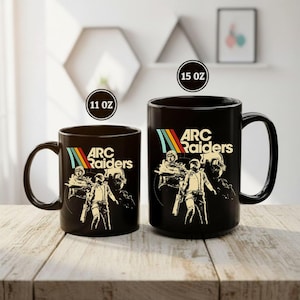 May include: Two black ceramic mugs with the text "ARC Raiders" and a retro graphic of figures in space suits. The mugs are labeled as 11 oz and 15 oz. The design features a rainbow stripe.