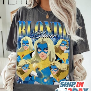 May include: Dark gray t-shirt featuring a graphic of a blonde superhero with a blue mask and yellow and blue costume. The text "BLONDE Blazer" is displayed in a blue and yellow font. The word "PEPPER" is at the top.