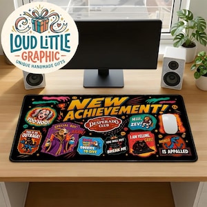 May include: A rectangular black desk mat featuring a colorful graphic print with the text NEW ACHIEVEMENT! and various cartoon illustrations including a goat, a woman, and several speech bubbles with phrases like THIS IS AN OUTRAGE and MONGO IS APPALLED.