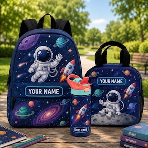 May include: A dark blue backpack, a matching lunch bag, and a water bottle, all featuring a space theme with astronauts, rockets, and planets. Each item includes a rectangular label with the text YOUR NAME printed in white letters.
