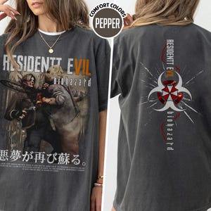 May include: Dark gray t-shirt featuring a graphic from the Resident Evil video game. The front displays the game title and characters, while the back has the biohazard symbol and game title. The shirt has short sleeves and a crew neck.