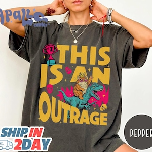 May include: Dark gray t-shirt with the text "THIS IS AN OUTRAGE" in yellow. The graphic features a cat wearing a crown and sunglasses riding a blue dinosaur. A trophy and skulls are also present.