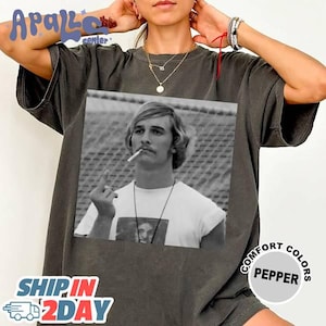 May include: Dark gray t-shirt featuring a black and white image of a man smoking a cigarette. The image is centered on the shirt. The shirt has the text "COMFORT COLORS PEPPER" on the right side. The shirt also has the text "SHIP IN 2DAY" on the bottom left.