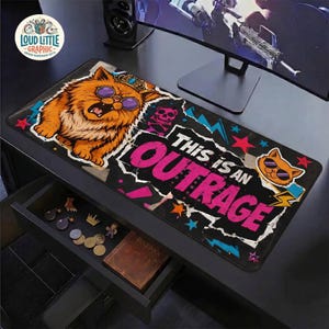 May include: A rectangular desk mat featuring a large orange cat wearing purple sunglasses and a crown. The design includes the text This is an OUTRAGE in bold pink letters, surrounded by colorful stars, lightning bolts, and a small skull icon.