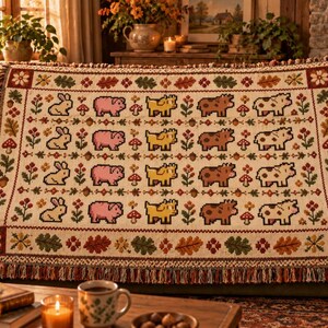 May include: A woven throw blanket featuring a repeating grid pattern of pixelated rabbits, pigs, yellow dogs, brown cows, and spotted cows, interspersed with mushrooms, acorns, and flowers, finished with a decorative fringed edge.