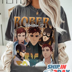 May include: Dark gray t-shirt featuring a graphic of a man with the text "ROBERT Robertson III". The graphic includes multiple illustrations of the same man in different poses and outfits. The shirt has a vintage, worn look.