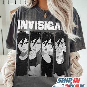 May include: Dark gray t-shirt featuring a graphic of four stylized faces with the word "INVISIGA" above them. The faces are in black and white, with varying expressions. The shirt has a vintage, worn look.