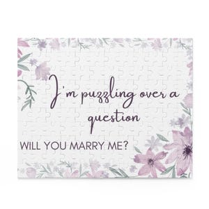 May include: A white jigsaw puzzle with a floral border. The puzzle features the text "I'm puzzling over a question. Will you marry me?" in purple lettering.