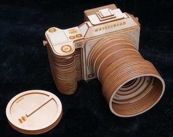 May include: A wooden camera model with the text "#Hasselblad" engraved on the top. The camera has a lens and a separate lens cap. The camera and cap are made of light brown wood and sit on a dark surface.