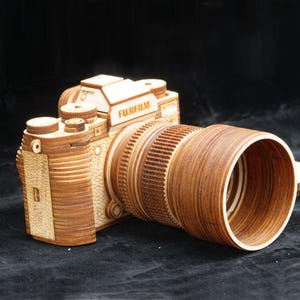 Puede incluir: Una cámara de madera con lente, con la marca "FUJIFILM". La cámara está hecha de madera marrón clara y oscura, con detalles intrincados en el cuerpo y la lente. El diseño imita un modelo de cámara clásico.