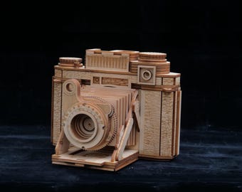 May include: A wooden model of a vintage camera, showcasing intricate details. The camera features a lens, dials, and a bellows design, all crafted from light-colored wood. The model is a detailed replica of a classic camera.