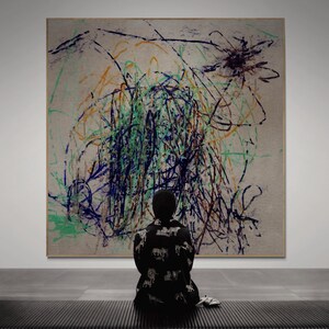May include: A large square canvas featuring abstract, chaotic scribbles in dark blue, bright green, and orange ink on a light beige background. The artwork is displayed on a wall, with a person wearing a black patterned jacket sitting in front.