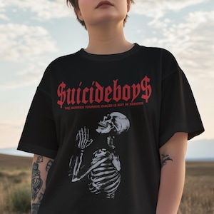 May include: Black t-shirt featuring the text "Suicideboys$" in red gothic font. Below the text is the phrase "THE NUMBER YOU HAVE DIALED IS NOT IN SERVICE". A white illustration of a skeleton praying is below the text.