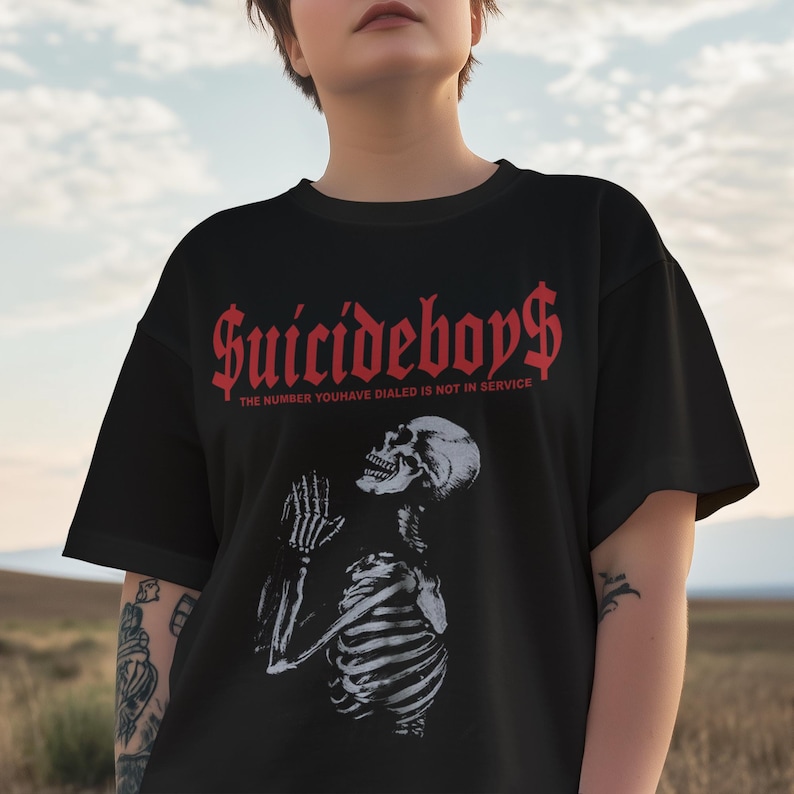 May include: Black t-shirt featuring the text "Suicideboys$" in red gothic font. Below the text is the phrase "THE NUMBER YOU HAVE DIALED IS NOT IN SERVICE". A white illustration of a skeleton praying is below the text.