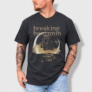 Op de afbeelding: Donkergrijs t-shirt met de tekst "breaking benjamin" in gouden letters. Het shirt heeft een cirkelvormige afbeelding met een Keltisch knoopontwerp en een goud- en bruintint.