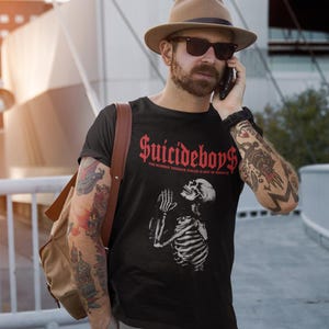 May include: Black t-shirt with the text "Suicideboys" in red gothic font and a skeleton graphic. The shirt also has the text "THE NUMBER YOU HAVE DIALED IS NOT IN SERVICE". The person is wearing a brown hat and sunglasses.