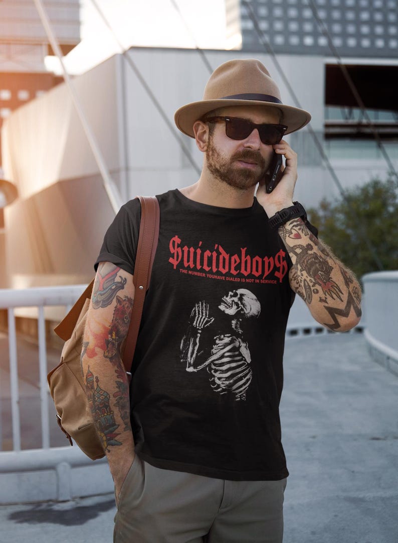 May include: Black t-shirt with the text "Suicideboys" in red gothic font and a skeleton graphic. The shirt also has the text "THE NUMBER YOU HAVE DIALED IS NOT IN SERVICE". The person is wearing a brown hat and sunglasses.