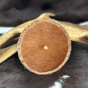 Op de afbeelding: Een ronde, bruine leren onderzetter met een gestikte rand. De onderzetter heeft een klein, rond gat in het midden.
