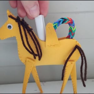 May include: A yellow felt horse with brown yarn mane, tail, and legs. The horse has a blue eye and a rainbow-colored rubber band loop on its back. A hand holds a white paper roll above the horse.