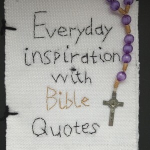 Könnte beinhalten: Ein weißes Stoffbuch mit den Worten "Everyday inspiration with Bible Quotes" in schwarzem und goldenem Faden gestickt. Ein lila Perlenrosenkranz mit einem silbernen Kruzifix liegt neben dem Buch.