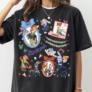 May include: Black t-shirt featuring colorful cartoon characters from Zootopia. The design includes images of a fox and a rabbit, along with the word "Zootopia" and the word "Bestie". The shirt has a relaxed fit.