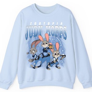 May include: Light blue sweatshirt featuring the text "ZOOTOPIA JUDY HOPPS" above a graphic of three cartoon rabbits in police uniforms. The rabbits are set against a backdrop of blue lightning bolts.