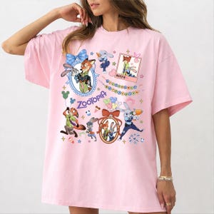 May include: A light pink t-shirt featuring a colorful graphic of characters from the animated movie Zootopia. The design includes images of a fox, a bunny, and other characters, with the word "Zootopia" prominently displayed.