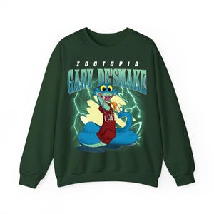 May include: Dark green sweatshirt featuring a cartoon snake character from Zootopia, named Gary De'Smake. The snake is blue with a red hoodie and is set against a background of lightning bolts. The word "Zootopia" and "Gary De'Smake" are printed on the sweatshirt.