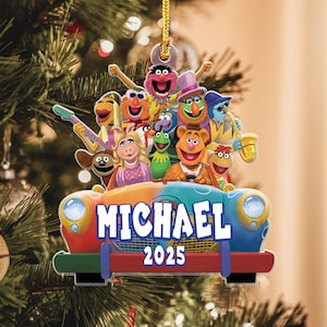 Könnte beinhalten: Ein farbenfroher Christbaumschmuck mit einer Gruppe von Muppet-Charakteren in einem Auto. Das Auto ist mehrfarbig mit dem Namen "MICHAEL" und dem Jahr "2025" bedruckt. Der Anhänger hängt an einer goldenen Schnur.