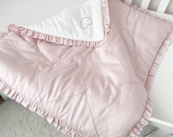 Puede incluir: Una manta de bebé acolchada de color rosa con un ribete de volantes blancos y un corazón dorado bordado en la parte superior.