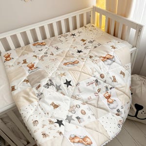 May include: A quilted baby blanket featuring a patchwork design with beige and white squares. The fabric is decorated with illustrations of brown teddy bears, black stars, toy cars, and building blocks, finished with a white pom-pom trim edge.