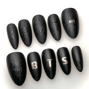 Może przedstawiać: Zestaw dziesięciu matowych, czarnych sztucznych paznokci. Każdy paznokieć ma unikalny wzór, w tym cyfrę "2", litery "BTS", błyskawicę i inne abstrakcyjne wzory. Paznokcie są owalne i mają gładkie, nierefleksyjne wykończenie.