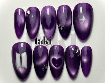 Könnte beinhalten: Ein Set aus zehn mandelförmigen Press-on-Nägeln mit einem tiefvioletten Katzenaugen-Finish. Das Design zeigt ein weißes BTS-Logo, einen vierzackigen Stern, eine Herzform und kleine klare Strasssteine. Der Text 'taki NAILS' ist mittig zu sehen.