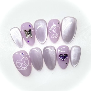 May include: A set of lavender-colored artificial nails with various designs. Some nails feature silver accents like a butterfly and hearts, while others have white line art. The nails are oval-shaped and have a glossy finish.