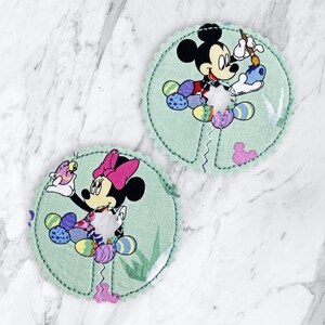 May include: Two green fabric circles with embroidered images of Mickey and Minnie Mouse. Mickey is holding a paintbrush and Minnie is holding a small bird. Both circles have a white center with a hole in the middle.