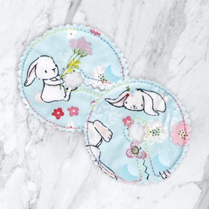 May include: Two light blue fabric nursing pads with a white bunny rabbit and floral print. The pads have a scalloped edge and a hole in the center.