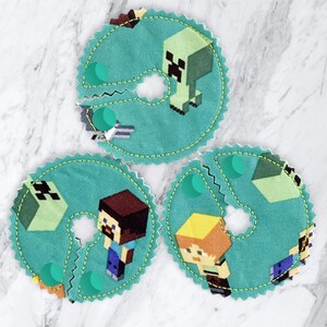 May include: Three teal fabric nursing pads with a white zig-zag stitch around the edges. Each pad features a different Minecraft character: a green creeper, a brown-haired character with a blue shirt, and a blonde character with a red shirt.