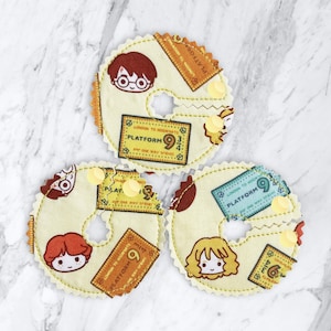 May include: Three yellow fabric circles with a hole in the center. Each circle has a different character from the Harry Potter series, including Harry Potter, Ron Weasley, and Hermione Granger. The circles also have a ticket design with the text "Platform 9 3/4" and "London to Hogwarts".